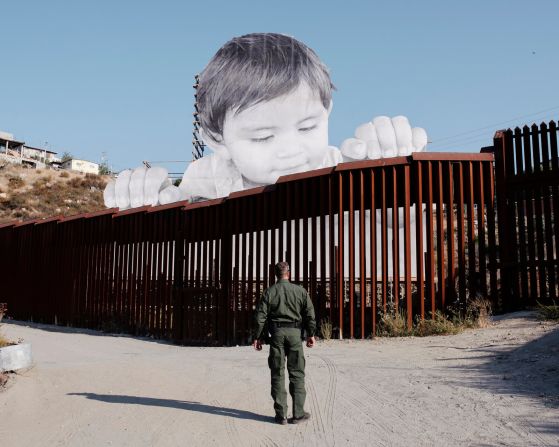 Un oficial de la Patrulla Fronteriza de EE. UU. se encuentra cerca de obras de arte instaladas en el lado mexicano de la frontera cerca de Tecate, California. La obra, basada en una foto de un niño mexicano, fue creada por el artista francés JR.