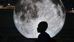 A boy stands near a model of the moon hanging above the Olympic swimming pool at the National Aquatics Center, known as the Water Cube, in Beijing on July 11, 2018. - The moon model is part of an exhibition about China's moon exploration technology and achievements. China landed its first rover on the moon in 2013 and plans to land a rover on the far side of the moon later in 2018. (Photo by GREG BAKER / AFP)