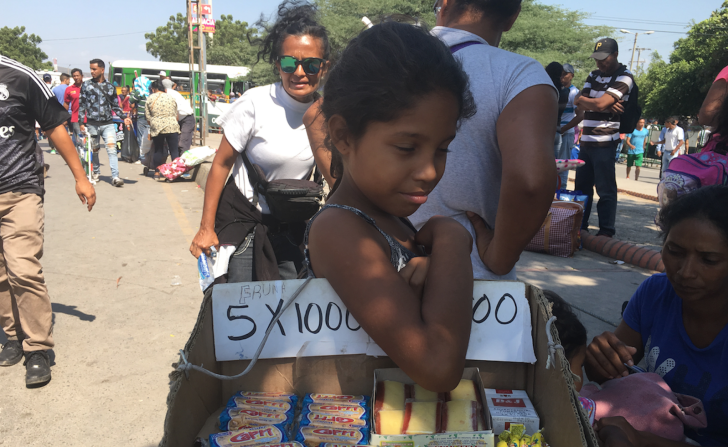 Una niña sostiene una caja con productos para la venta como galletas, bocadillos y cigarrillos.