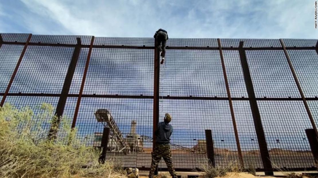 Un migrante de Ecuador visto trepando por la cerca del muro fronterizo entre Estados Unidos y México mientras un traficante de personas sostiene la escalera abajo.