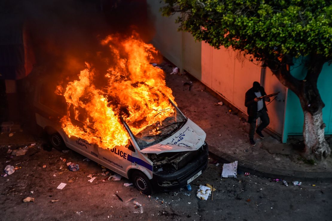 Un hombre con el rostro cubierto sostiene un arma y camina junto a un vehículo policial en llamas durante los enfrentamientos entre manifestantes argelinos y la policía antidisturbios en Argel, capital de Argelia.