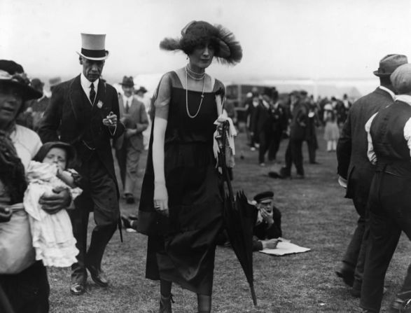 Una asistente a Royal Ascot 1922 camina bajo la lluvia con un vestido suelto y collar de perlas, dos íconos de la moda de la década.