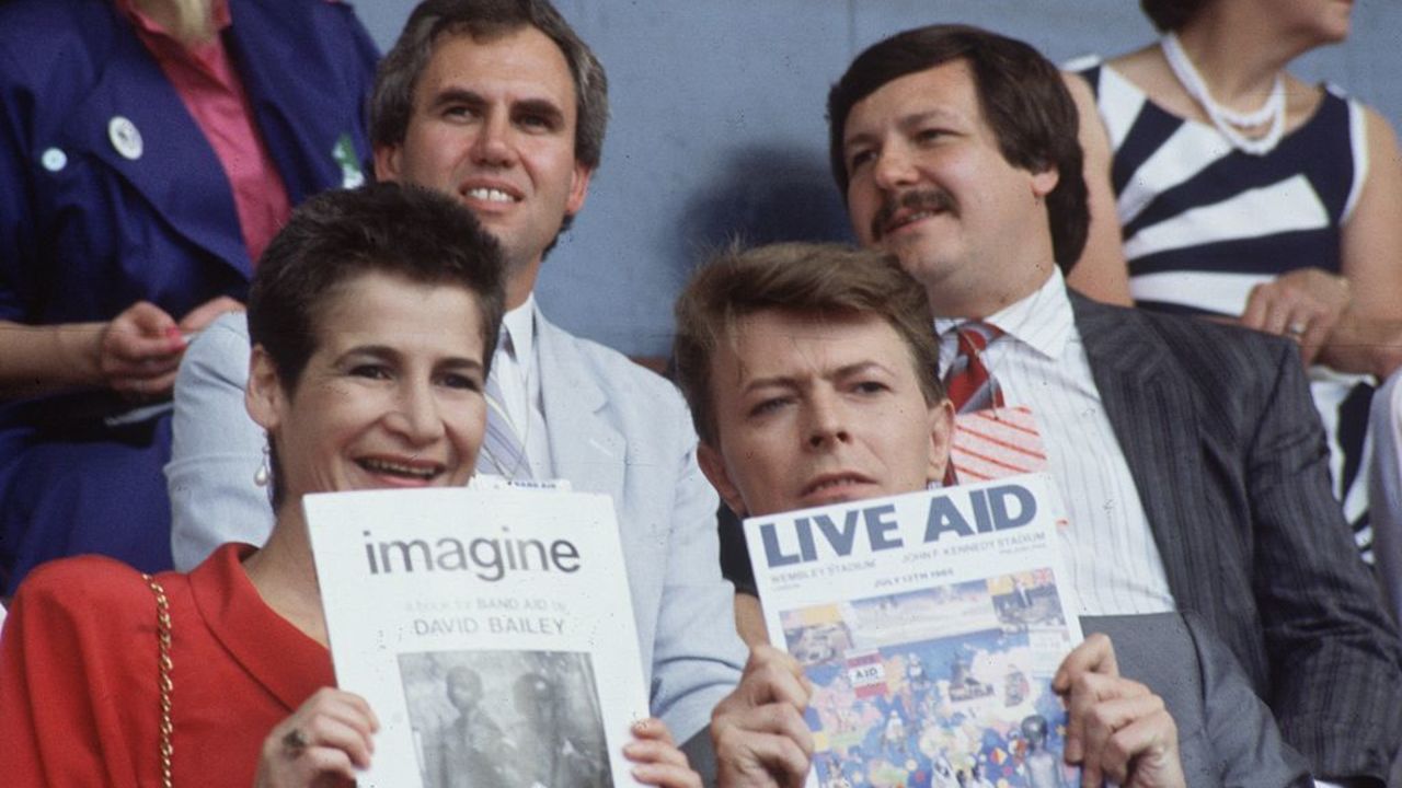 13th July 1985:  Singer David Bowie (David Jones) with a friend at the 'Live Aid' concert in Wembley Stadium, London. David Bowie is holding up a copy of the Live Aid programme.