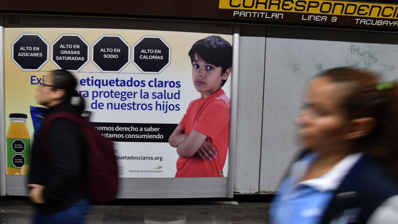 Women walk past a sign that refers to the labeling of products, in the subway of Mexico City, on October 01, 2019. - The Mexican Congress discusses Tuesday changes in the labeling of "junk food", such as to those products which have high content of sugar or saturated fats, with the aim of reducing the high rate of obesity in the population, although the industry opposes the measure. (Photo by RODRIGO ARANGUA / AFP)