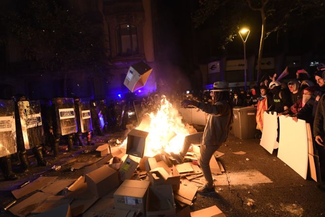 Policías y manifestantes de pie a ambos lados de una barricada de cajas en llamas.