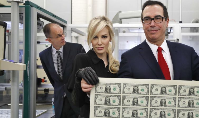 El secretario del Tesoro de Estados Unidos, Steven Mnuchin, y su esposa, Louise Linton, sostienen una hoja de billetes nuevos de 1 dólar en noviembre de 2017. La serie era la primera en presentar la firma de Mnuchin. Linton fue ampliamente criticada a principios de año cuando publicó una foto en Instagram que la mostraba bajando de un avión del gobierno y presumiendo de su exclusivo vestuario al mencionar a los diseñadores en la leyenda de la imagen. La fotografía provocó una investigación sobre el uso de Mnuchin de aviones del gobierno. El inspector general del Departamento del Tesoro finalmente no encontró evidencia de irregularidades, pero dijo que la administración Trump había acortado procesos durante el procedimiento de aprobación de los viajes de Mnuchin.