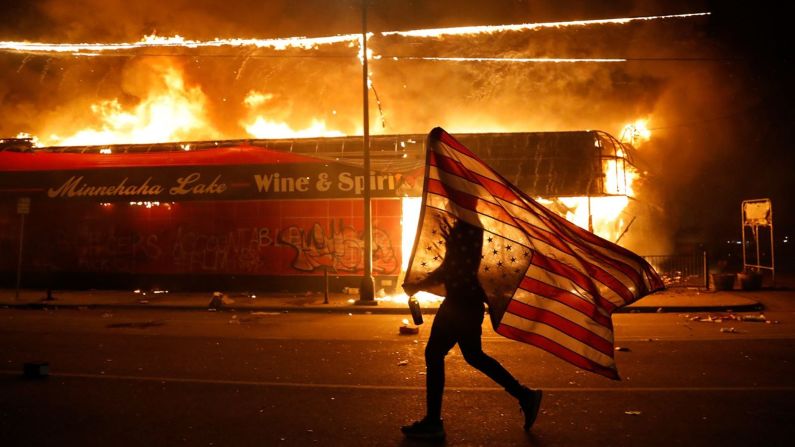 Un manifestante carga una bandera de Estados Unidos frente a una tienda de licores en llamas en Minneapolis este jueves.