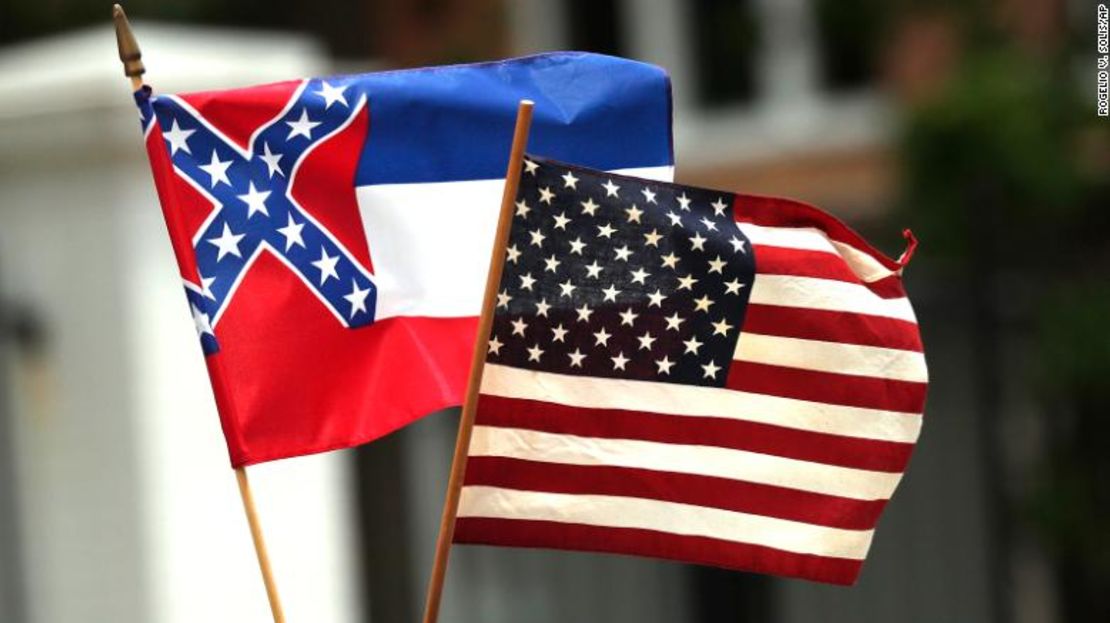 En esta fotografía, del 25 de abril de 2020, un participante sostiene una pequeña bandera del estado de Mississippi durante una protesta de "reapertura de Mississippi" al pasar junto a la Mansión del Gobernador, en el fondo, en la ciudad de Jackson. La bandera actual tiene en la parte del estandarte el diseño de la bandera de batalla confederada de la era de la Guerra Civil, que ha sido el centro de un debate a fuego lento sobre su eliminación o reemplazo.