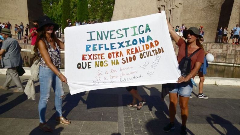 Dos mujeres sostienen una pancarta que dice "Investiga, reflexiona, existe otra realidad que nos ha sido ocultada".