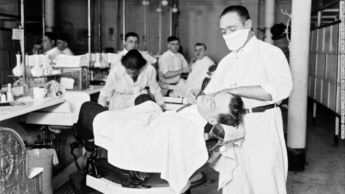 En una barbería no especificada, un hombre recibe un afeitado de un barbero con una máscara contra la influenza durante la pandemia en curso, Chicago, Illinois, 1918.