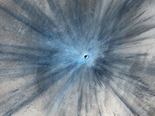 Un cráter de impacto reciente y drástico domina esta imagen tomada por la cámara HiRISE en noviembre de 2013. El cráter se extiende aproximadamente 30,5 metros y está rodeado por una gran zona de explosión de rayos. Debido a que el terreno donde se formó el cráter es polvoriento, el cráter reciente aparece azul en el color realzado de la imagen, debido a la eliminación del polvo rojizo en esa área.