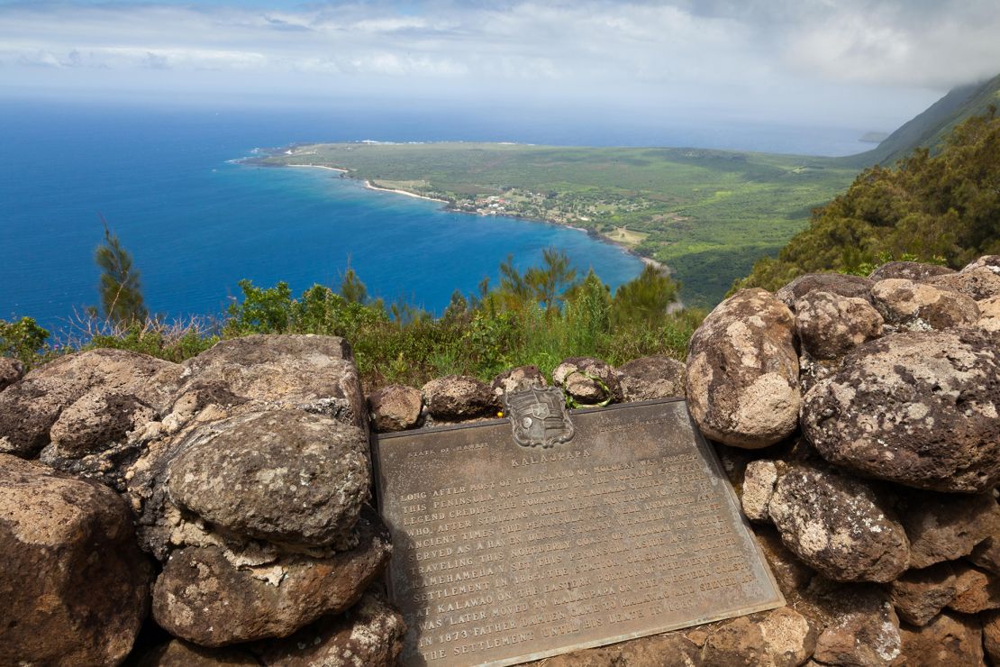 A plaque explains some of the area's history at Kalaupapa Lookout. The peninsula emerges at the base of towering sea cliffs.