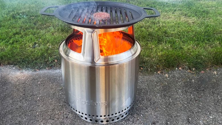 A burger cooks on a grill over a bright orange flame. The fire is inside a stainless steel fire pit on a backyard patio with green grass in the background.