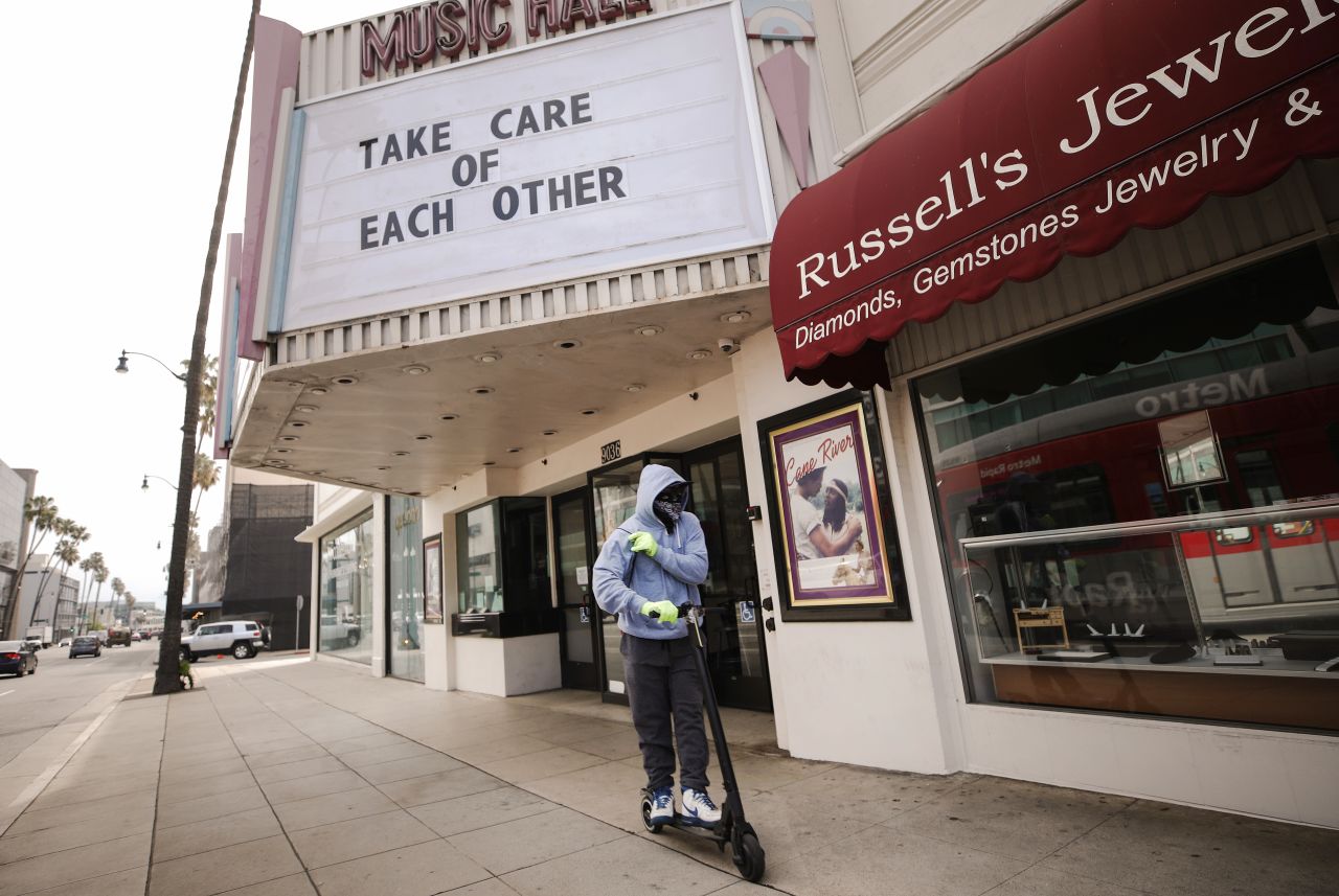 A man wears gloves and a bandanna across his face while riding a scooter past a shuttered movie theater, with the message "Take Care of Each Other" displayed on the marquee, on March 18, in Beverly Hills, California. 