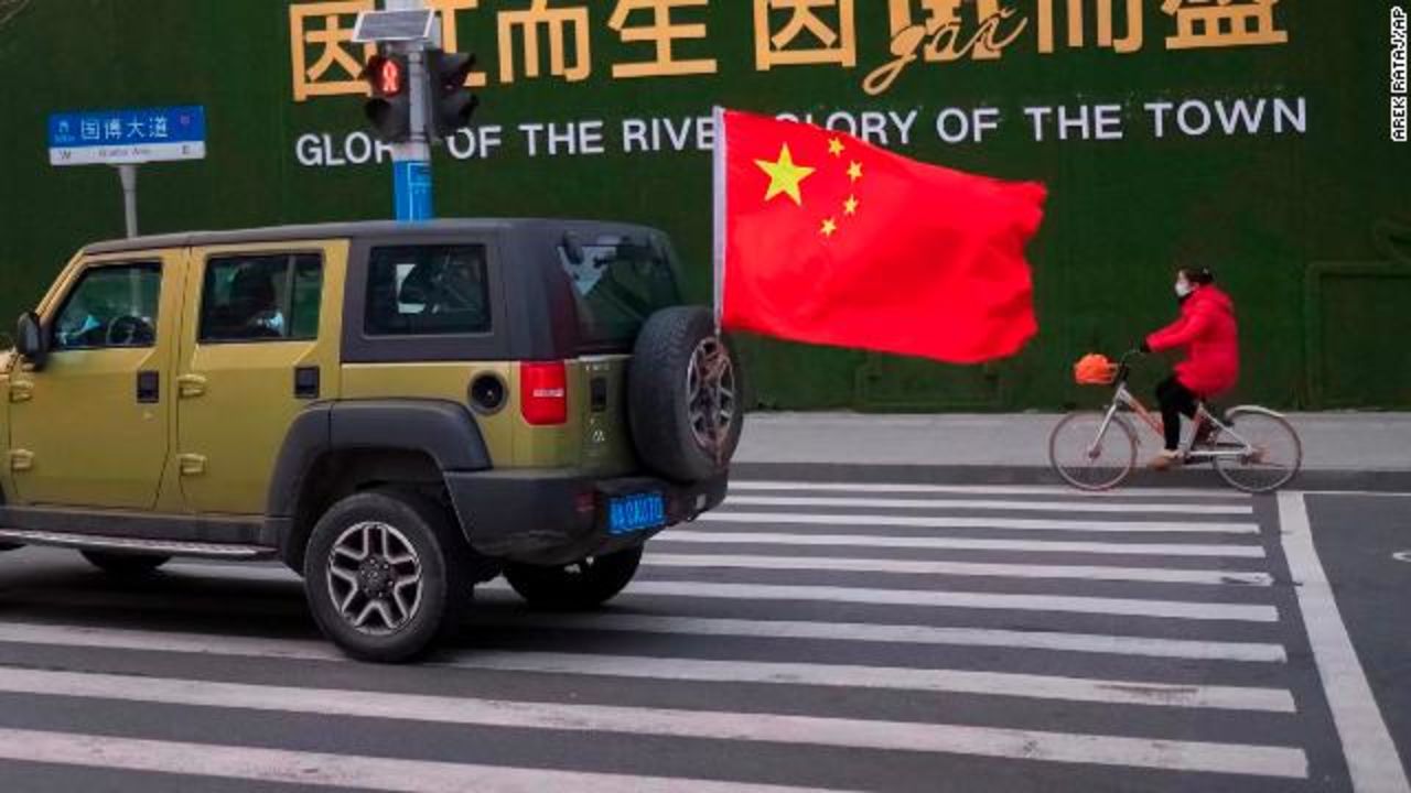 A vehicle flying a Chinese flag drives past a woman wearing a face mask as she rides a bicycle along a street in Wuhan.