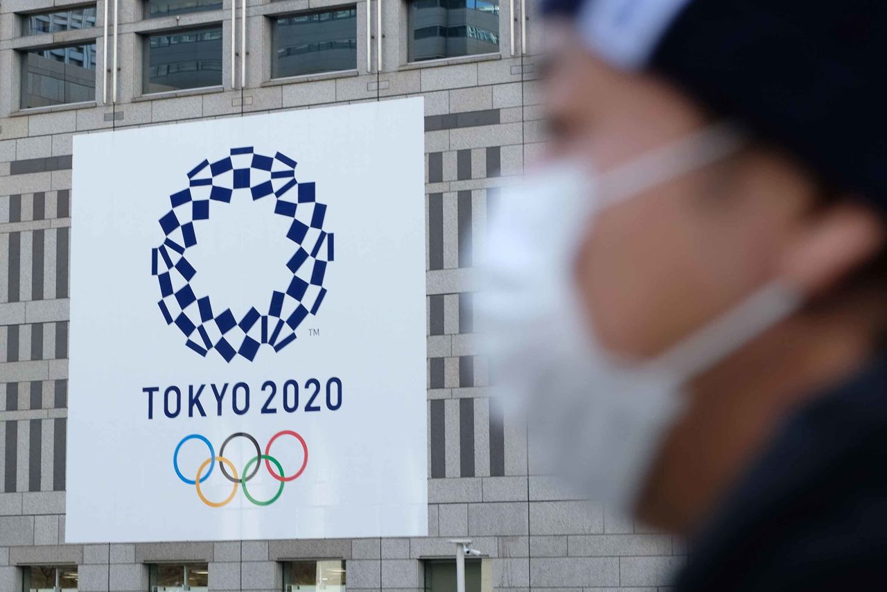 A man wearing a mask passes the logo of the Tokyo 2020 Olympic Games displayed on the Tokyo Metropolitan Government building on March 19, in Tokyo, Japan.