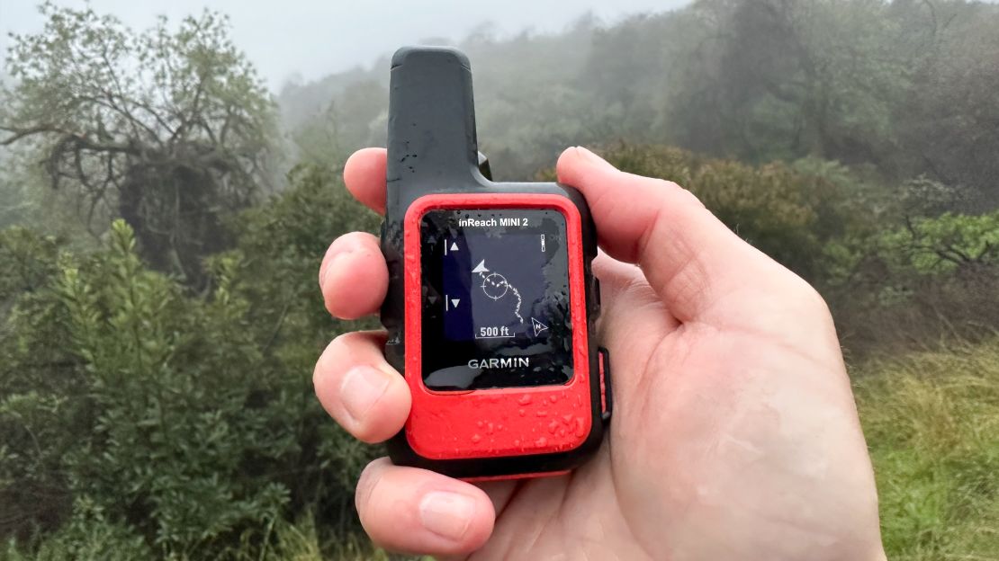 The Garmin inReach Mini 2 is held in a right hand with a forested area in the background.