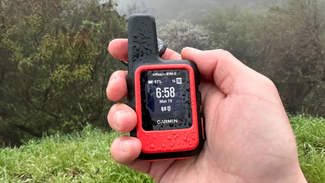 The Garmin inReach Mini 2 is held in a right hand while displaying the time with a forested view in the background.