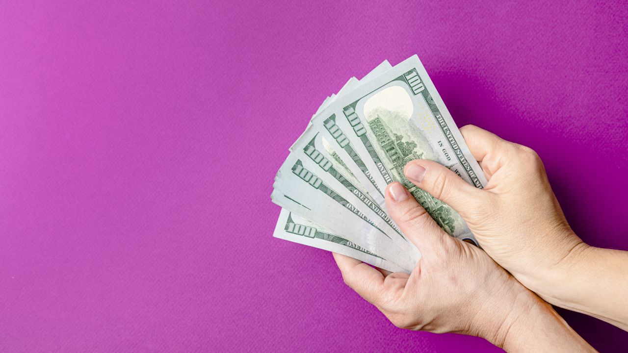 Hands holding multiple $100 bills over a pink background.