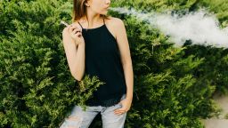 Trendy girl exhaling a cloud of vapor while smoking an electronic cigarette, standing near the bushes, looking away and keeping the hand in the pocket. Outdoors.
