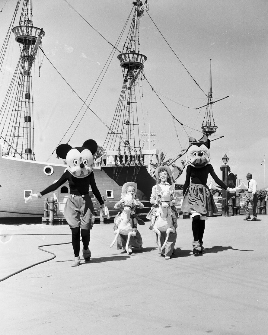 Disney icons Mickey and Minnie Mouse walk with children near a pirate ship on Disneyland's opening day.