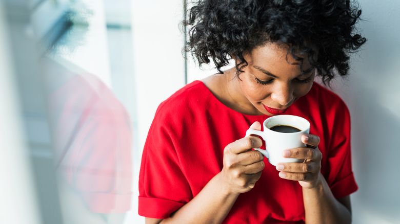 A close-up of a woman standing by the window holding a cup of coffee, smelling it. Copy space.