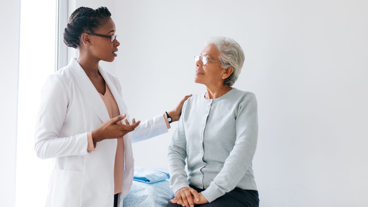 A black female doctor standing face to face with a senior female patient and talking to her in doctor's office.