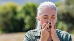 Senior woman having flu using nasal spray to help herself. Woman using a nasal spray at park for allergy. Ill mature woman with grey hair inhaling medicine against allergy.