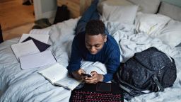 High angle view of distracted teenage boy using mobile phone while lying on bed by books at home