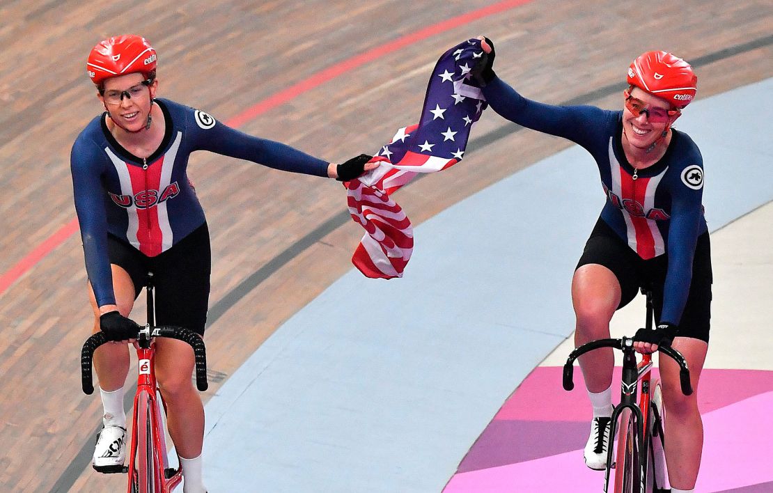 Two women smiles while riding bicycles, holding an American flag between them. 