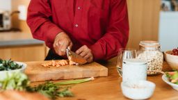 Midsection of retired man chopping carrot on cutting board. Senior male is preparing vegan meal at kitchen island. He is cooking in kitchen at home.