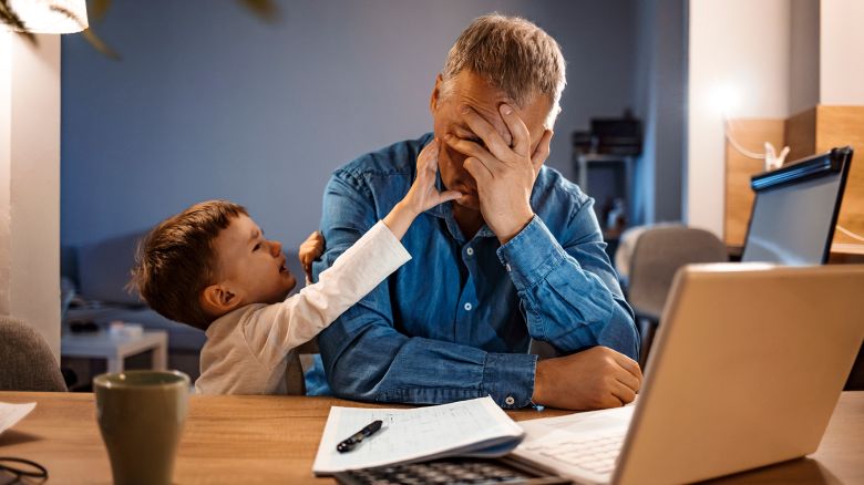 Busy mature businessman working from home and watching his crying son at night, who is is yelling at him. Upset displaced father forced to work from home as the pandemic corona virus (COVID-19) forces many employees to work from home. Real people. Copy space.