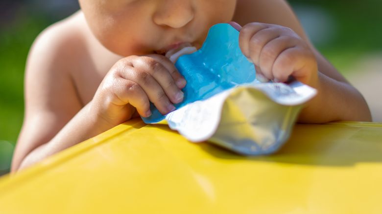 baby eating fruit puree in pouch and looking into the food in front of the yellow table. on the background is a green garden on a sunny day in blur