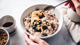 Close-up of a woman mixing oats flour, banana and blueberries in a bowl. Female making healthy breakfast in kitchen.