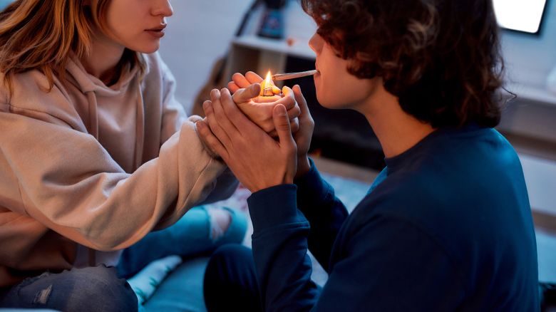 Young girl lighting cannabis cigarette for her boyfriend while sitting on the couch at home. Young couple smoking marijuana joint and relaxing. Marijuana, drugs, cannabis and weed legalization