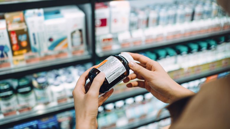 Over the shoulder view of young Asian woman browsing through medical products and reading the label on a bottle of medicine in front of the shelves in a pharmacy