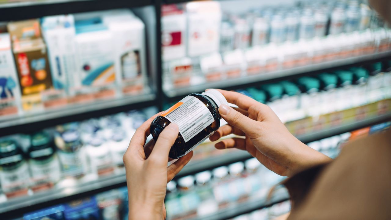 Over the shoulder view of young Asian woman browsing through medical products and reading the label on a bottle of medicine in front of the shelves in a pharmacy