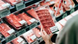 Over the shoulder view of young Asian woman shopping in a supermarket. She is choosing meat and holding a packet of organic beef in front of the refrigerated section.