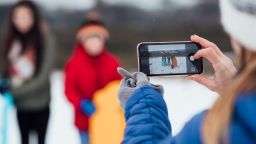 An unrecognisable caucasian woman taking a photo of her two children on her smart phone. They are wearing warm winter clothing and accessories in a snowy, rural, outdoor setting.