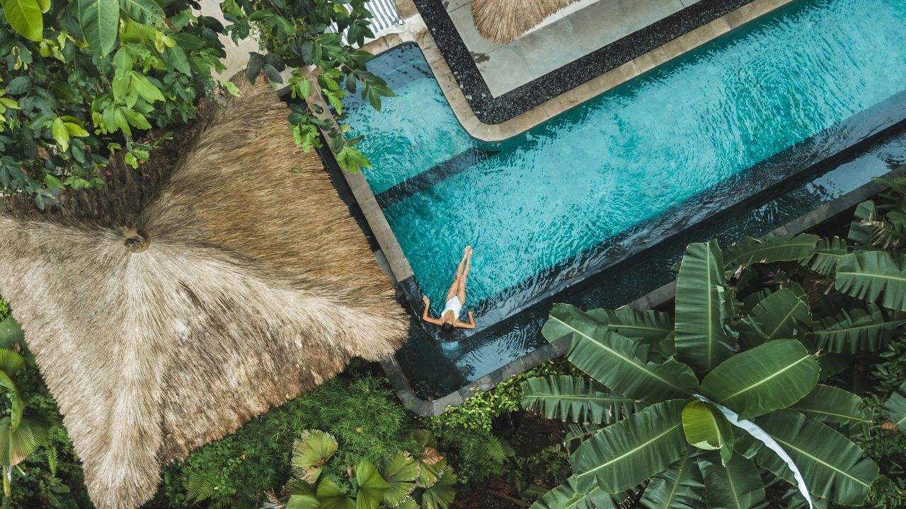 Girl relaxation in private swimming pool hiding in jungle. Wealth travel. Top view, drone point of view directly from above. Vacations in Bali. Tropical and exotic asian nature. Wellness and vitality. Balinese traditional architecture, hotel with straw roof.