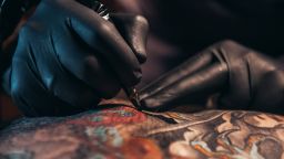 Hands of a tattoo artist wearing black gloves and holding a machine while creating a picture on a man back and the ink is dripping