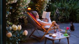 Woman relaxing on deck chair in back yard, reading a book with her cat watching - stock photo