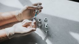 A hand wearing latex gloves hold a phial of clear liquid and a syringe. The medical professional uses the syringe to extract the medicine from the glass bottle. Conceptual with space for copy.