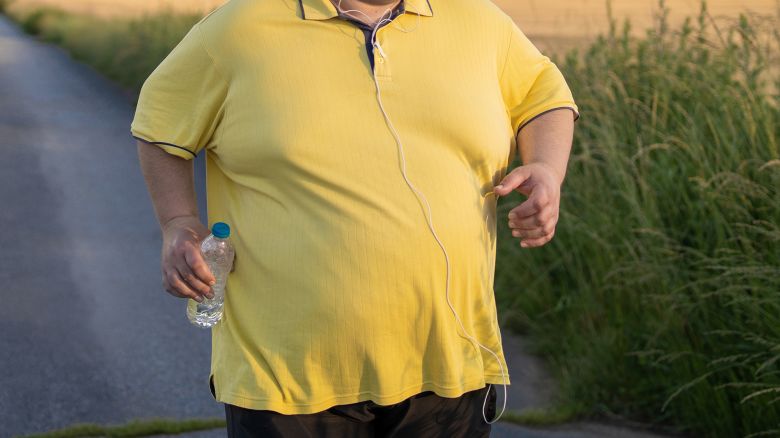 A large man listening to some music while he jogs down an empty country road, wearing comfortable clothing and a headband he carries a water bottle