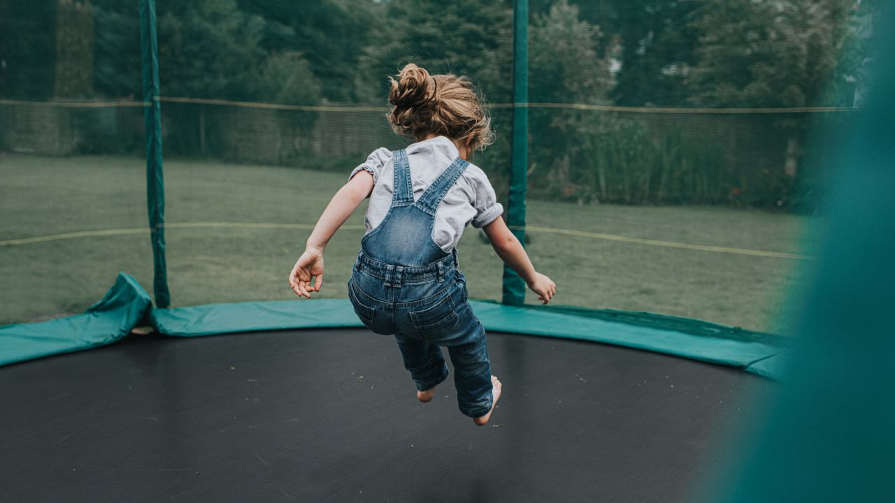 A little girl bounces on a large trampoline in a domestic garden. She is in mid air, surrounded by safety netting.