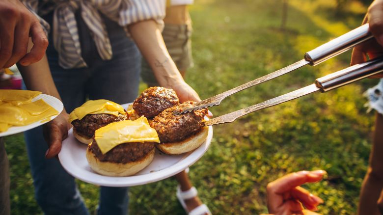 Cheese. Image of tasty homemade burger with melting cheese, tomatoes and grilled meat. Group of friends having nice outdoor picnic, making food, drinking beer and having fun.