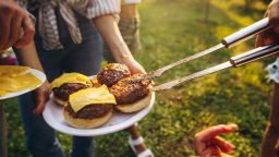 Cheese. Image of tasty homemade burger with melting cheese, tomatoes and grilled meat. Group of friends having nice outdoor picnic, making food, drinking beer and having fun.