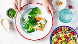 Children's hands eating fish with creamy sauce and broccoli in white plate, top view. Baby food concept.