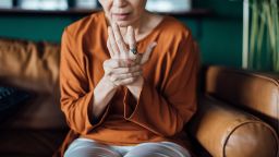 Senior Asian woman rubbing her hands in discomfort, suffering from arthritis in her hand while sitting on sofa at home. Elderly and health issues concept