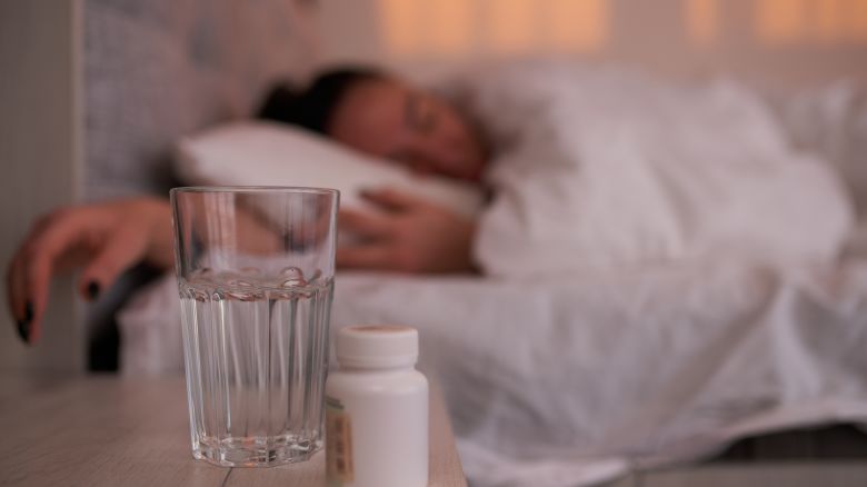 Soft focus of jar with sleeping pills and glass of water placed on bedside table near sleeping female in morning at home.